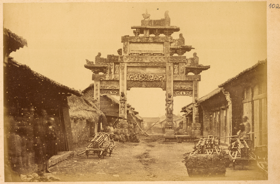 Ornamental gateway (pailou) from the Han dynasty (202 BCE – 220 CE) across a street lined with small shops. Hanzhong, Shaanxi Province, China (1875).