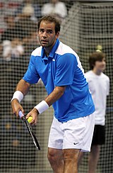 Fotografía en color de un hombre que sostiene una raqueta de tenis y una bola en cada mano. Él se está preparando para servir.