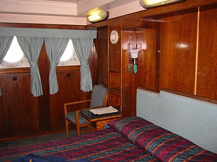 First Class accommodation on Queen Mary, converted into a present-day hotel room with modern curtains, bedding, fixtures and amenities surrounded by original wood panelling and portholes