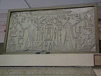 Kamenná stéla visící na zdi, na které se nacházejí dva polonazí muži s vlčí hlavou a sukní, jeden otočen na levou stranu a druhý na pravou. Oba mají pozvednuté levé ruce, muž na levé straně na levou stranu a muž na pravé straně na pravou stranu a drží v nich anchy. Druhá ruka obou mužů taktéž drží anch, ale není pozvednutá. Kolem mužů se nachází hieroglyfy a symboly