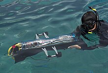 A male student holds a small vehicle in a body of water. The vehicle is enclosed in a clear plastic tube and the machinery inside is clearly visible.