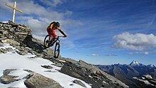 VTTiste descendant un chemin légèrement enneigé le long d'une montagne. À sa gauche, une croix a été installée au sommet de la montagne.