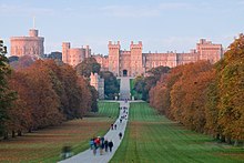 Photographie d'un château de couleur rouge à l'horizon. Une route empruntée par des promeneurs et située au milieu d'une percée dans une foret mène jusqu'au château.