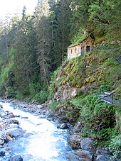 Cliché numérique couleur. Premier plan avec un torrent. En arrière, flanc de montagne avec une forêt de sapins et et une habitation perchée.