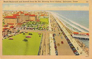 A postcard shows a wide oceanside boulevard lined with cars and pedestrians. A large hotel complex lies on the opposite site from the water, as well as parks and other buildings.