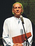 A photograph of Gheorghe Zamfir. Standing in front of a microphone, he wears a white shirt and holds a nai panflute in his hands.