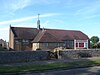 Side view of a long, barn-like brick building with an off-centre extension on the near side, above which is a star-shaped metal spire on top of a metal crown-like device. Small rectangular windows are placed at regular intervals. A cobbled wall, bench and grass verge are in the foreground.