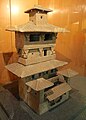 Side view of a Han pottery tower model with a mid-floor balcony and a courtyard gatehouse flanked by smaller towers; the dougong support brackets are clearly visible.