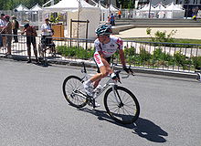 Jalan balap sepeda di jersey putih dengan garis merah di atas putih sepeda, melihat ke belakang dia keluar dari frame. Penonton yang terlihat di pinggir jalan di belakang barikade.