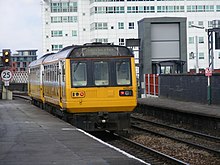 An orange painted train with two carriages leaving a railway platform.