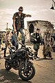 Zdjęcie przedstawia mężczyznę przebranego w strój postapokaliptyczny. Stoi on na motorze i wskazuje palcem do przodu. W tle zdjęcia przechadzają się cztery osoby wśród ruin lotniska Kluczewo.