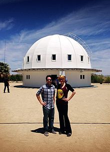 Man with light skin and dark hair and woman with light hair and light skin stand in front of a white building with a domed roof