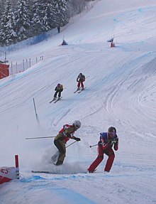 Vier Skifahrer in einer Abfahrt, zwei im Vordergrund, losgelöst von den beiden anderen.