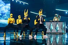 Foto von zwei Personen in gelben Masken und schwarzem Anzug sowie drei ebenfalls in gelb-schwarz gekleideten Tänzern auf einer Bühne