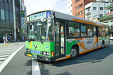 Długi zielony autobus niskopodłogowy na pierwszym planie, tuż za nim ulica i budynki w mieście.