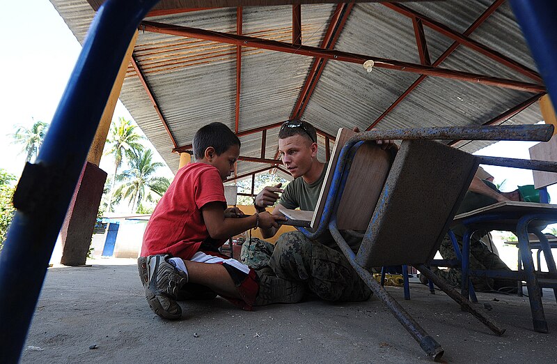 File:US Navy 110128-N-7589W-405 Cpl. Paul Fuit, assigned to 2nd Marine Logistics Group, shows a child how to use a rasp to smooth wood desks at Escuela.jpg