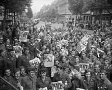 Multitud de jubilosos soldados aliados en una calle;  varios sostienen un periódico con la palabra Piece escrita en letras grandes.