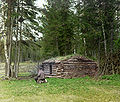 Woodcutter sitting next to log cabin. 1912