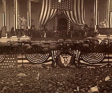 Fotografia de uma plataforma decorada com bandeiras americanas.  Muitos participantes estão segurando guarda-chuvas e todos na multidão abaixo estão usando chapéus.