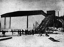 Photographie d'un entrepôt devant un lac gelé sur lequel travaille un groupe d'hommes. Un tapis roulant situé à côté du bâtiment permet de faire monter la glace à son sommet.