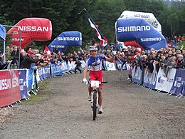 Homme sur un VTT portant une tenue bleu-blanc-rouge et tenant un drapeau français dans la main gauche avec un public en arrière-plan.