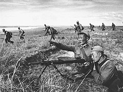 Photo en noir et blanc de soldats qui marchent, au premier plan deux soldats tirent à la mitrailleuse.