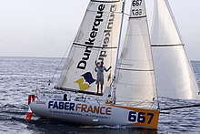 Le voilier 667, blanc et orangé, floqué « Dunkerque » et « Faber France ». Vu de profil, il avance sur l'océan. Le navigateur est debout sur la bôme, adossé à la grand-voile.