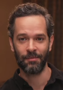 44 year-old man with short black hair and a beard smirking to the left.