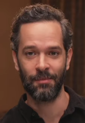 44 year-old man with short black hair and a beard smirking to the left of the camera.