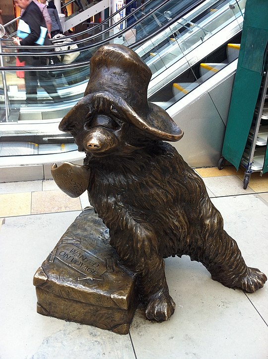 A bronze statue of Paddington Bear. Paddington wears a hat, has a label around his neck and sits on a suitcase.