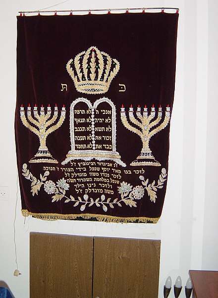 Cover of the ark of the Torah in the synagogue in Moshav Tsofit, Israel: Pictured in the center are the Tablets of the Ten Commandments; to their right and left are the 7-branched menorahs that were used in the Temple; above is the crown of the Torah