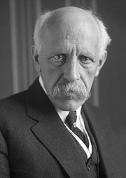 Un homme photographié en buste : chauve, épaisse moustache tombante, veste, gilet, chemise et cravate.
