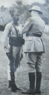 Foto av en svart soldat som står i oppmerksomhet, med de Gaulle sett bakfra, i en kolonihjelm og fester en dekorasjon på ham