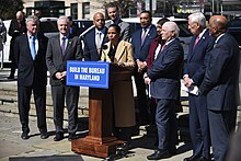 Alsobrooks stands and speaks at a podium with a sign saying "Build the Bureau in Maryland". She is surrounded by Democratic members from Maryland's congressional delegation and other statewide officials.