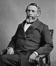 Photographie d'un homme avec une barbe poivre et sel en costume assis dans un fauteuil