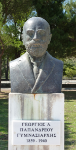 Photograph of a bust of a man with a bald head and a beard and mustache, on a white marble pillar with a plaque containing Greek writing and 1859 - 1940.