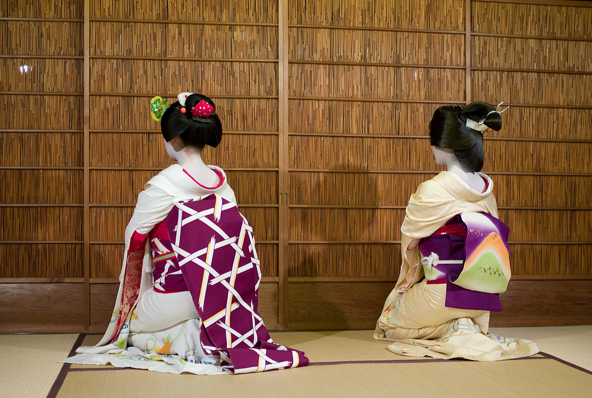 A maiko (on the left) and a geisha (on the right) facing away from the camera， sat on a tatami mat.