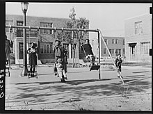 Eine Gruppe von ungefähr vier schwarzen Kindern spielt 1938 auf einer Schaukel, die von Gebäuden umgeben ist.