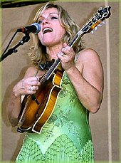 Une femme blonde vêtue d'une robe verte chantant et jouant de la mandoline.