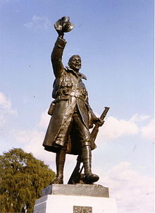 Patung prajurit dengan senapan di sisi kirinya dan mengangkat topi tinggi-tinggi di tangan kanannya.