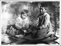Two Havasupai Indian children, the daughters of Chickapanagie's, enjoying a melon, ca. 1900. Both are wearing checkered dresses and are barefooted. They sit on a blanket with a zig-zag pattern with their hands in a half melon between them. Their hair is cut very short.