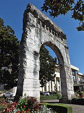 Photographie en couleurs des vestiges d'un arc de triomphe romain ; gra, d bâtiment à l'arrière-plan.
