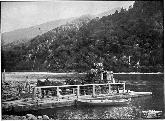 A flat boat with overhead guide wires, loaded with horses tied in front of a passenger coach up against the left-hand side of a river, with sloped bush-cover hills on the right-hand side