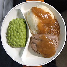 Fotografía de una placa de plástico moldeada con dos compartimentos;  en el más pequeño, un material verde que se asemeja a los guisantes en salsa, en el grande una bola más grande de comida que se asemeja al puré de papas o al arroz, parcialmente cubierta con una salsa marrón pegajosa que también cubre la bola contigua de comida, que se asemeja a un puré de hígado.