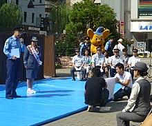 Two men (right, kneeling) hold cue cards to enable a public speech in Japan, 2016 Jaguar Yokota cuecards - shimbashi sep 1 2016.jpg