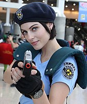 A woman wearing a costume at a convention from the waist up. She is wearing a dark blue beret, light blue shirt with shoulder armor and black gloves. She is pointing a pistol towards the viewer