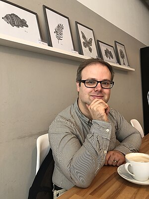 Matthew Toogood sitting at wooden table facing camera, his nearest facing hand is on his chin, there is a coffee in front of him, behind on the wall are photos standing on a white ledge.