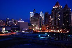 A very dark foreground fronts streets intersecting at a shallow diagonal. Buildings on the far side of the street are dark, with their interior lights visible. A dark blue sky is in the background.