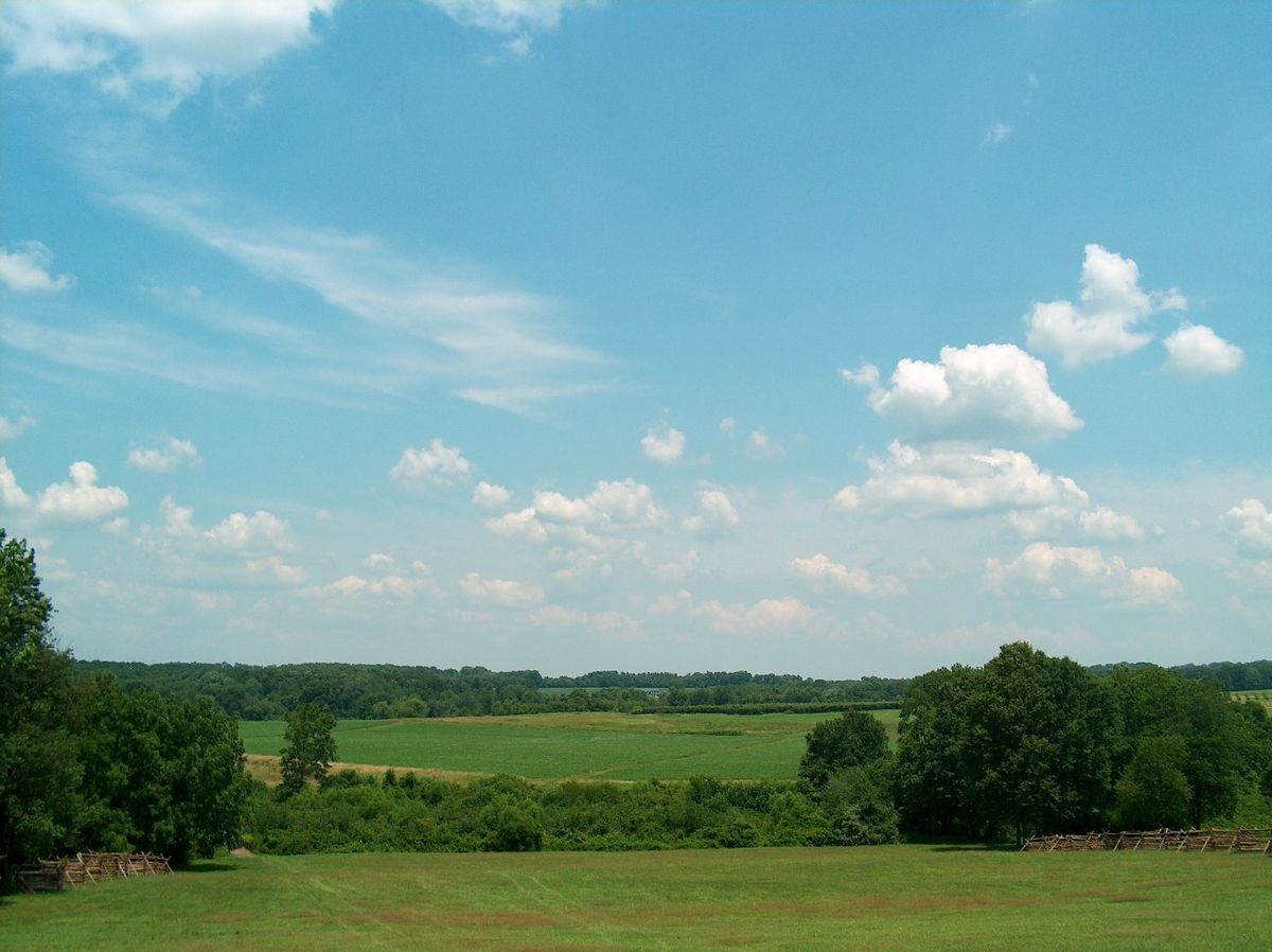 Photo shows a rural scene under partly cloudy blue skies. In the foreground a grassy hillside slopes down to a tree-filled ravine. On the other side are more open fields， with trees in the distance.