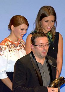 Photographie au format portrait et en couleurs de deux jeunes femmes souriant sur un podium, derrière un homme tenant une récompense et s'exprimant au micro.